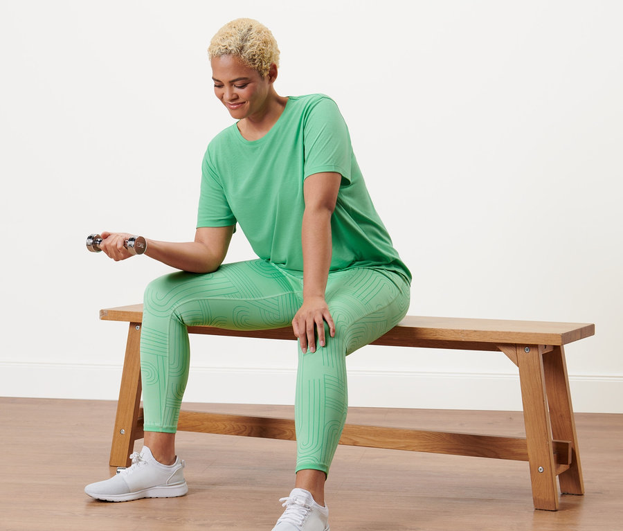 Femme assise sur un banc faisant des exercices d'haltères en portant un t-shirt de sport vert et un legging de sport réversible.