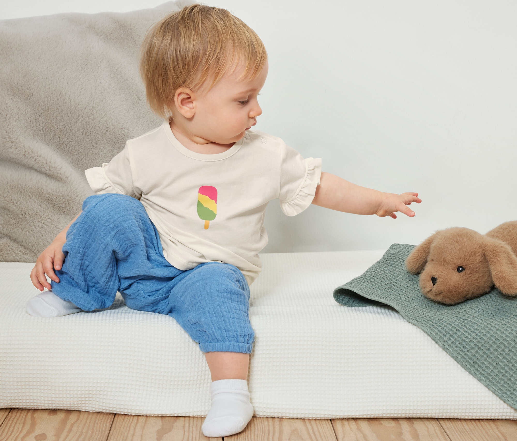 Un bébé est assis sur un lit et tend la main vers un chien en peluche. Il porte un t-shirt avec un dessin de glace. Un coussin gris est visible en arrière-plan. 3 t-shirts pour bébé sont présents.