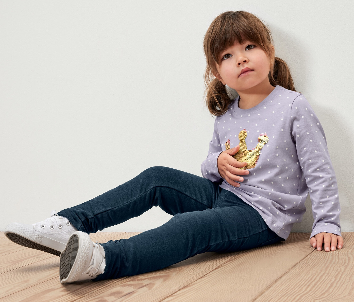 Une fillette est assise sur un plancher en bois et porte 2 jeggings pour enfant – Fit « Mia », une chemise violette avec une couronne et des baskets blanches.
