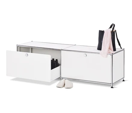Un meuble bas en métal blanc « CN3 » ouvert avec des tiroirs et des chaussures à l'intérieur, à côté duquel se trouve une paire de chaussons crème, un sac à main noir et une écharpe rose sur le dessus du meuble.
