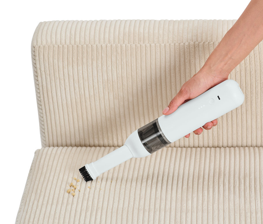 Une main aspire des miettes sur une chaise en tissu beige avec un aspirateur à main à batterie.