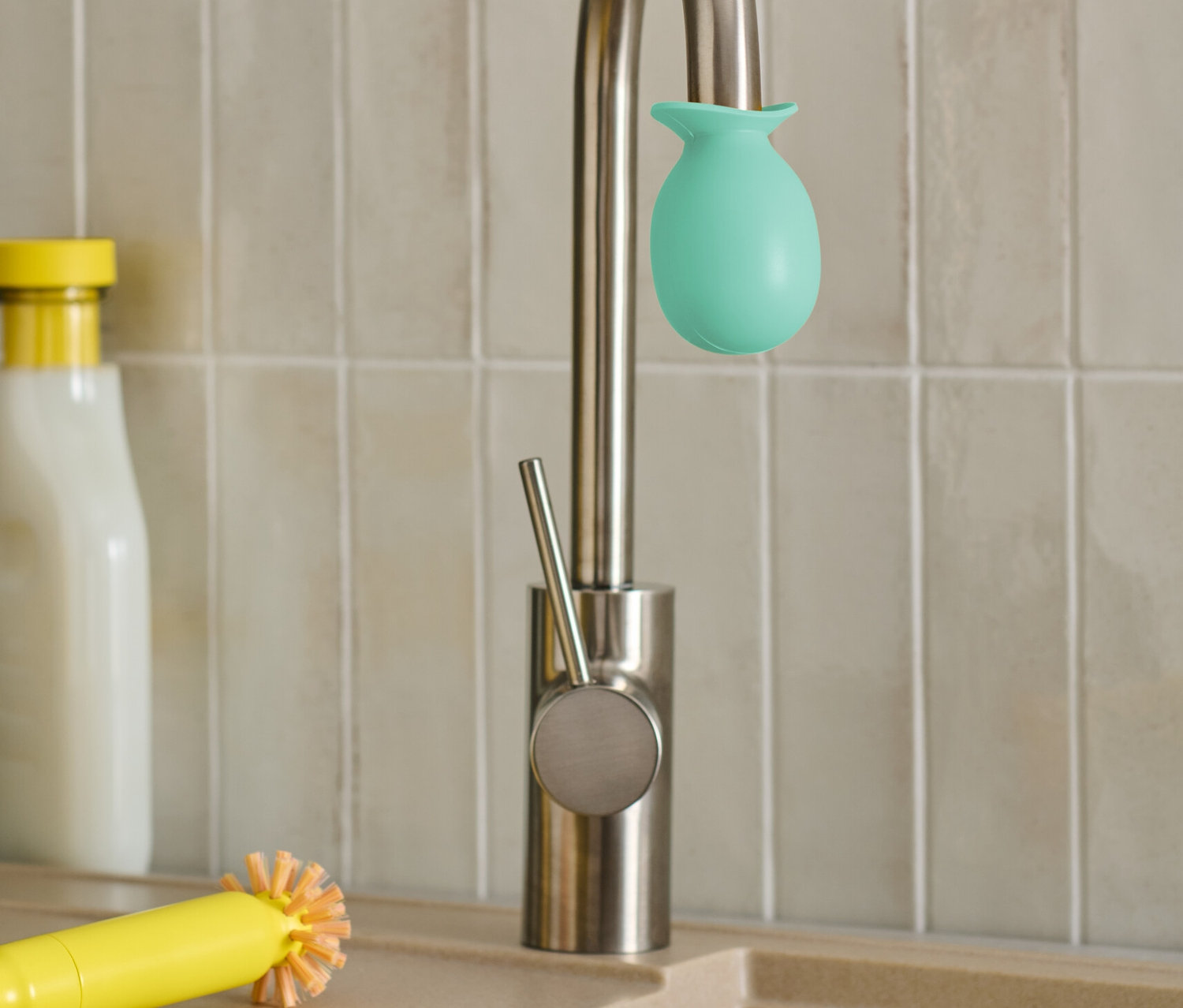 Un embout anti-calcaire pour robinet vert est fixé sur un robinet. À côté se trouvent une brosse jaune et un chiffon.