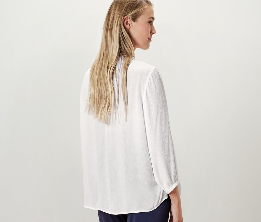 Une femme blonde pose dos à la caméra et porte un chemisier-tunique blanc.