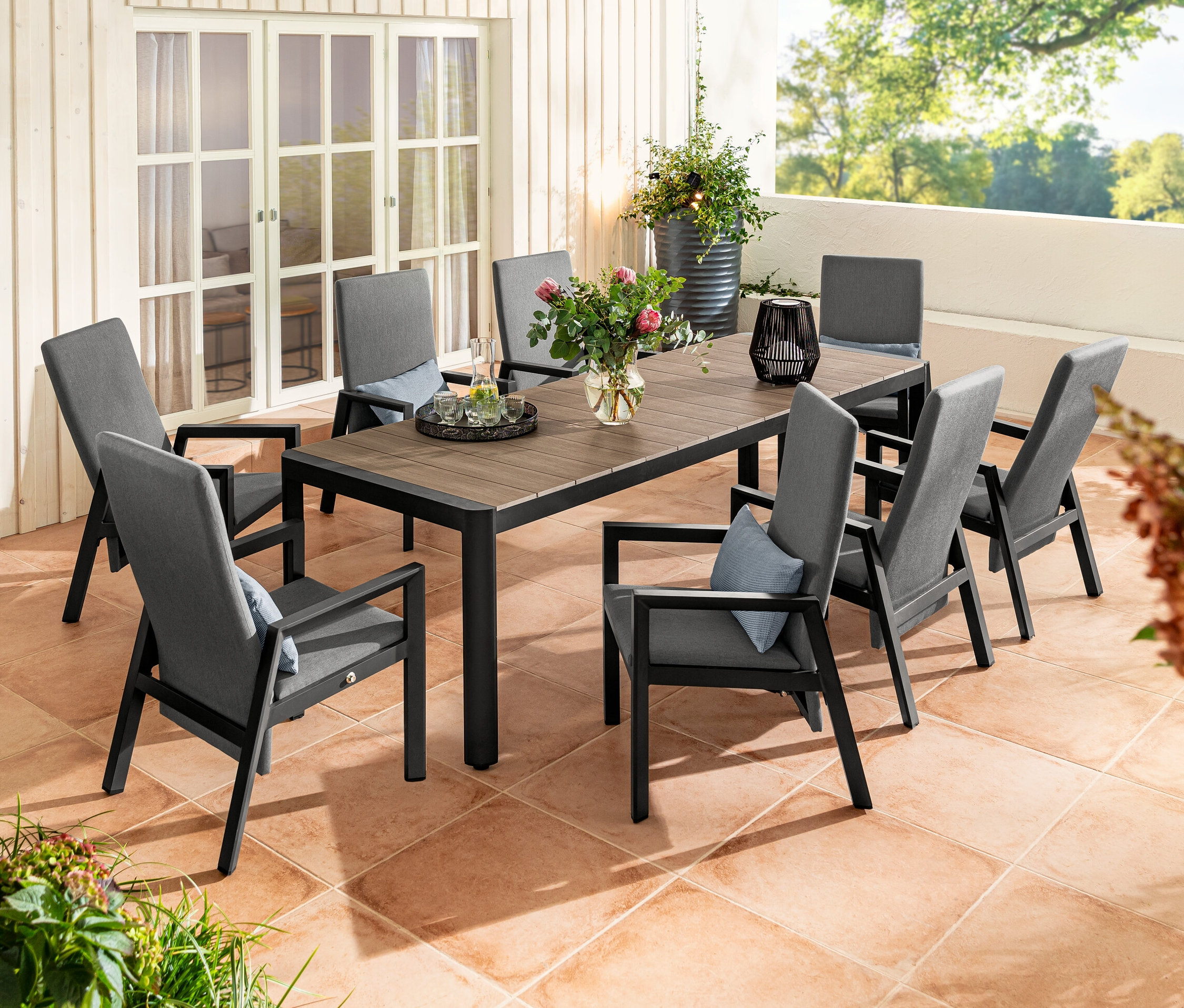 Une terrasse extérieure avec une table à rallonge avec plateau en polywood et huit fauteuils à haut dossier. Sur la table, il y a une lanterne solaire avec ruban en tissu et deux spots solaires.