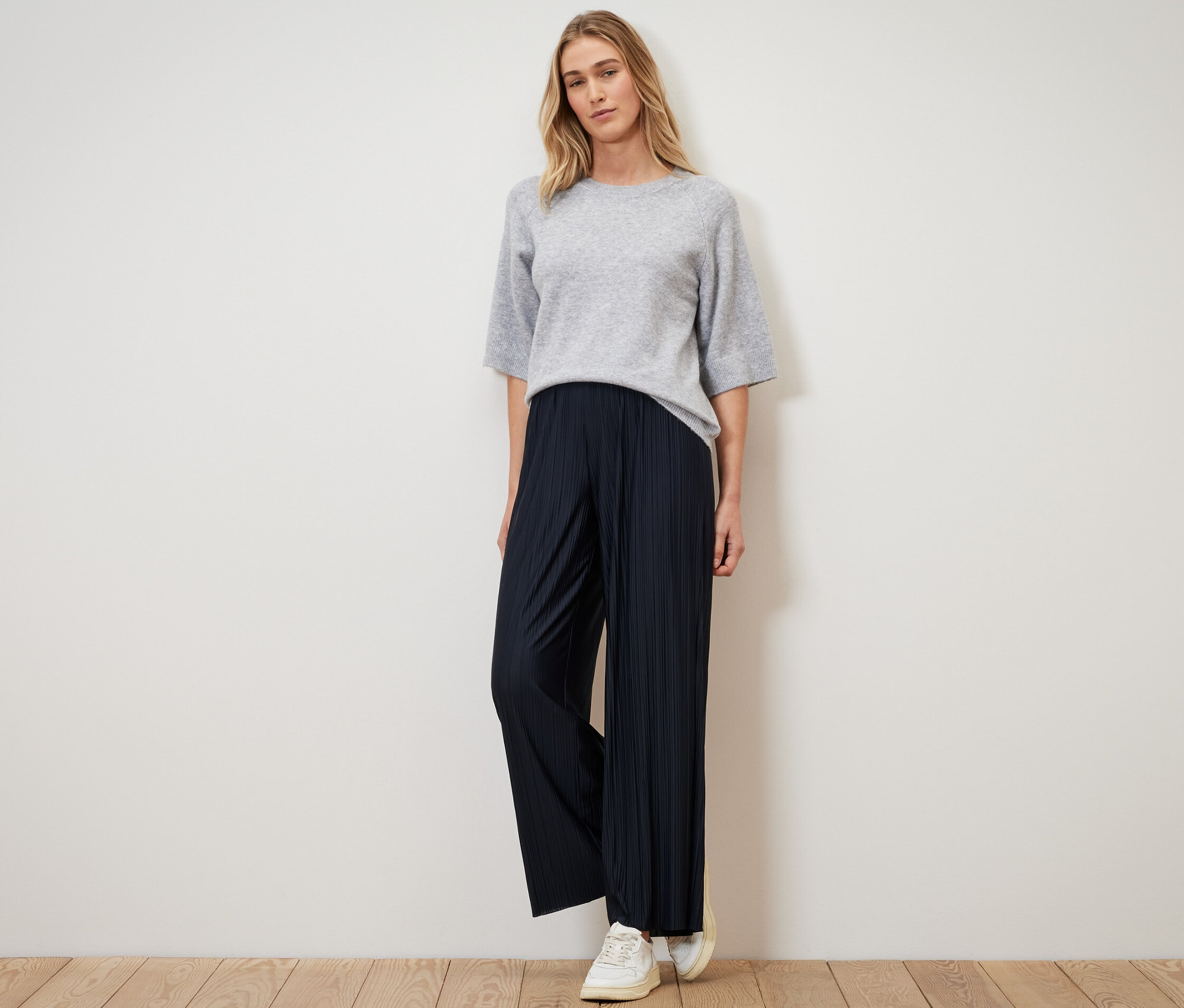 Une femme blonde pose en pull gris et pantalon plissé bleu foncé.