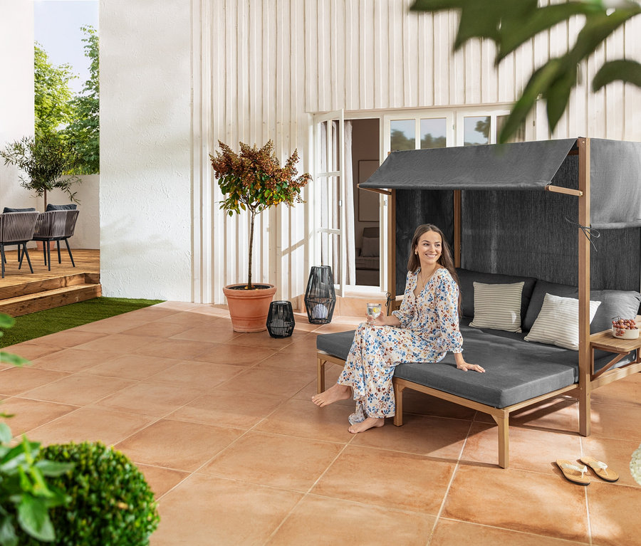Une femme est assise sur une chaise de plage Lounge « Tinus » avec repose-pieds extensible. À l'arrière-plan, il y a un fauteuil pour table à manger avec tressage textile et une table à rallonge « Elin ».