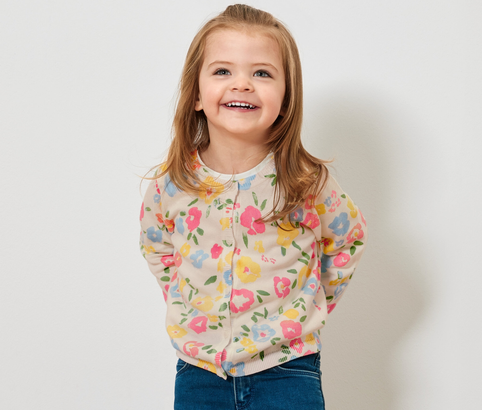 Une fillette souriante porte une jaquette en maille pour enfant à fleurs et un jean.
