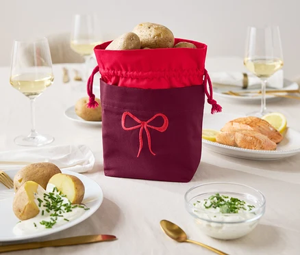 Sur une table, un sac en tissu bordeaux à bordure rouge, rempli de pommes de terre, à côté d'assiettes de pommes de terre avec de la sauce et de saumon avec du citron.
