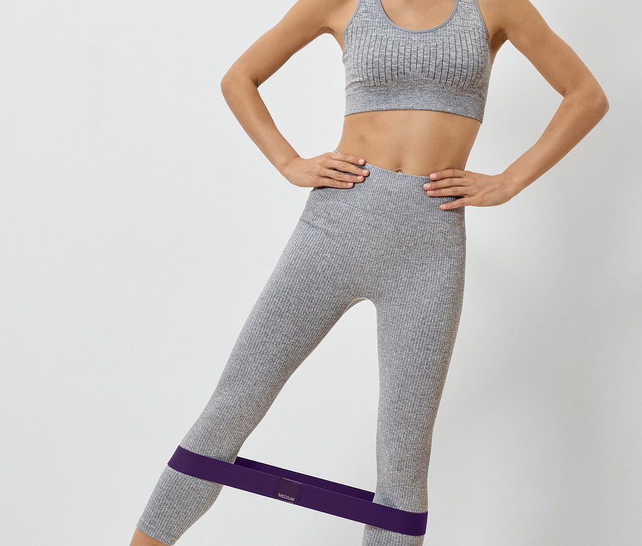 Femme en tenue de sport grise avec un élastique de fitness violet autour des jambes faisant de l'exercice.