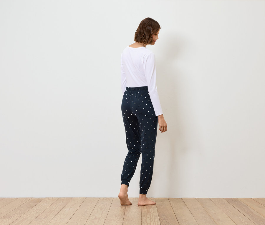 Une femme se tient dos à la caméra, portant un haut blanc à manches longues et un pantalon de jogging bleu marine à motifs.