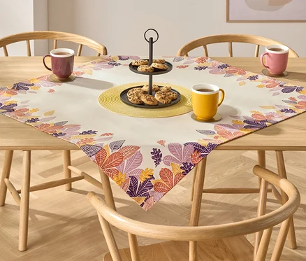 Sur une table en bois, une nappe aux motifs de feuilles d'automne. Sur la nappe, un présentoir à gâteaux avec des biscuits et trois tasses avec des boissons.