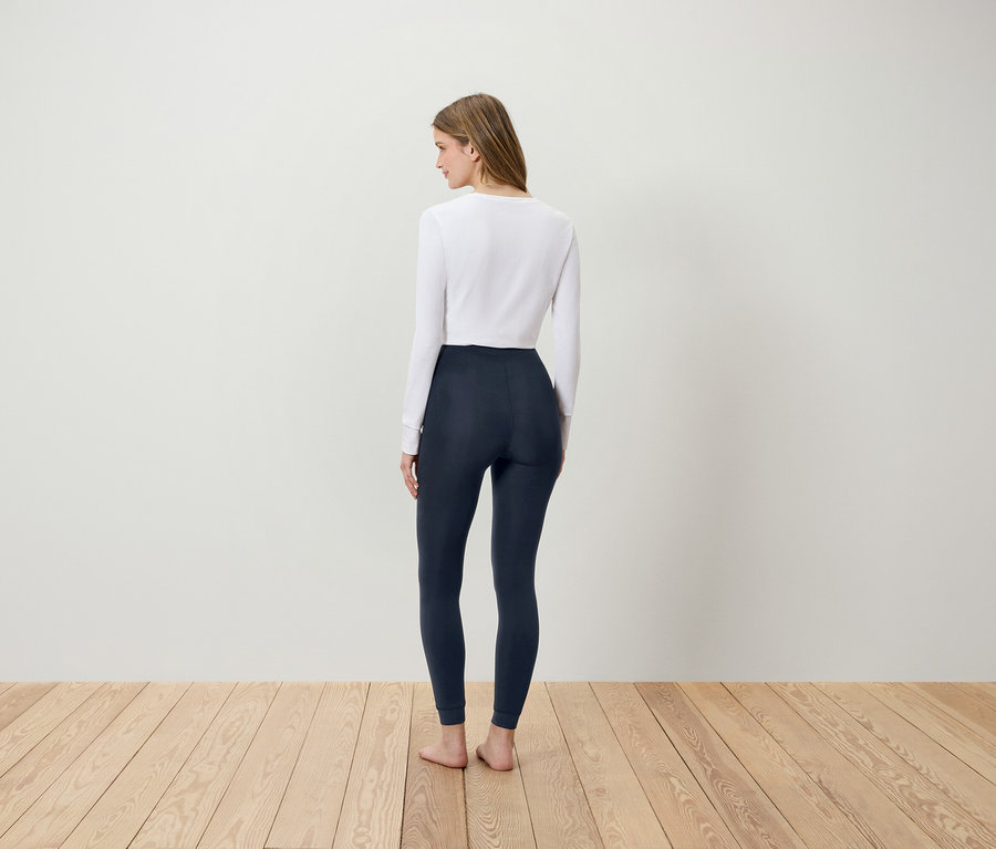 Une femme portant un T-shirt blanc et un legging bleu foncé, dos à la caméra, se tient sur un plancher en bois.