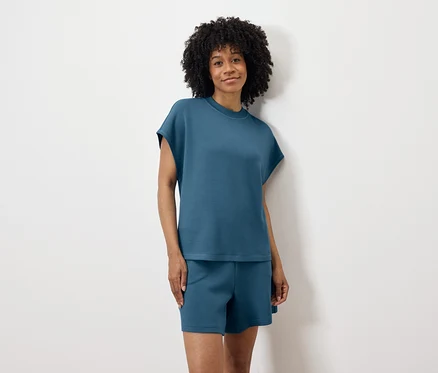Une femme aux cheveux bouclés, portant un t-shirt bleu et un short, se tient devant un mur blanc.