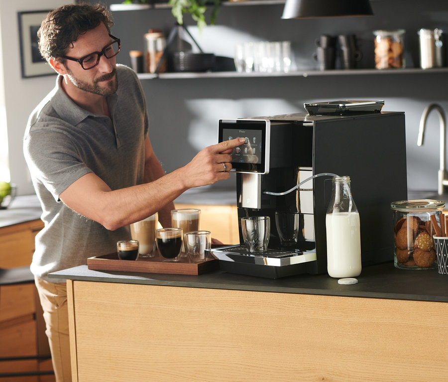 Un homme utilise une cafetière automatique Tchibo Office, noire. Des tasses à café et du lait sont posés sur la table.