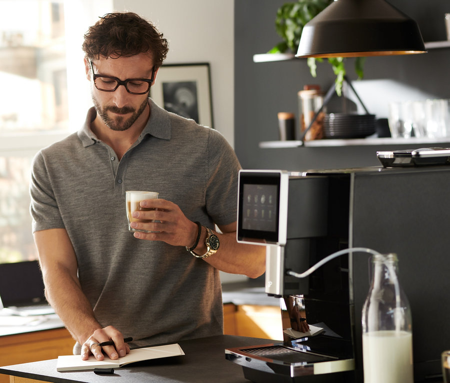 Un homme boit du café à côté de la machine à café »Tchibo Office« noire.