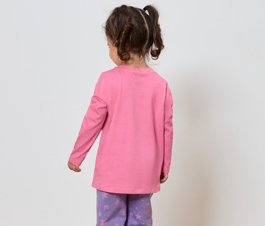 Petite fille aux cheveux bruns, vêtue d'un T-shirt rose à manches longues et d'un pantalon violet à motifs étoilés, se tient dos au spectateur.