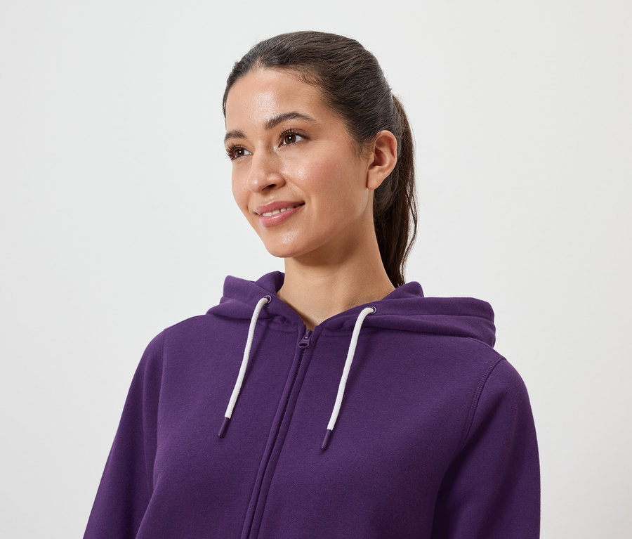 Une femme aux cheveux bruns coiffés en queue de cheval porte un sweat à capuche violet à fermeture éclair.