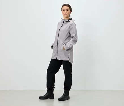 Une femme se tient debout, les mains dans les poches d'une veste à capuche grise, un pantalon noir et des bottes noires.