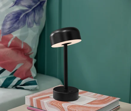Une lampe de table à LED noire sans fil est posée sur un livre, avec un coussin à fleurs en arrière-plan.