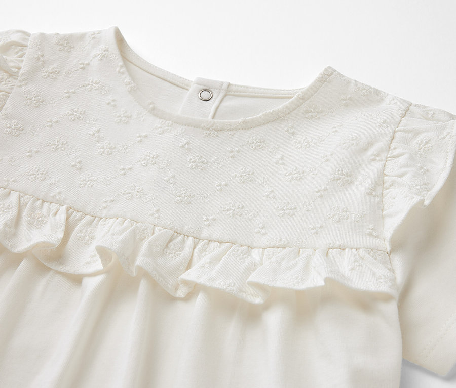 Gros plan sur un t-shirt enfant crème à manches courtes avec des volants décoratifs et des broderies sur la poitrine.