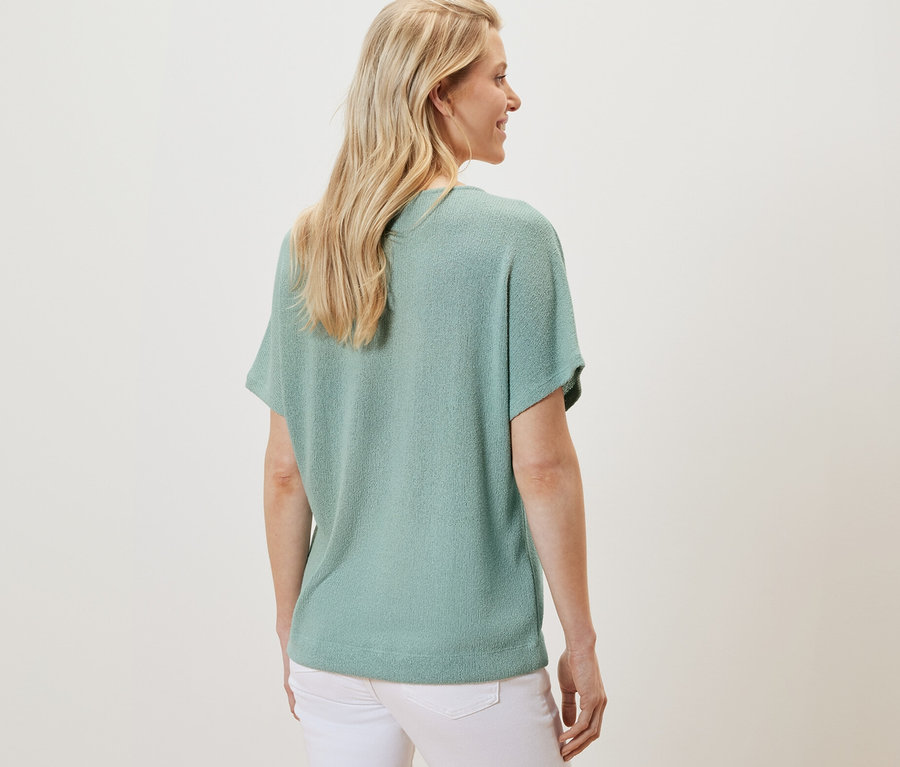 Vue arrière d'une femme blonde portant un t-shirt structuré sauge et un pantalon blanc.