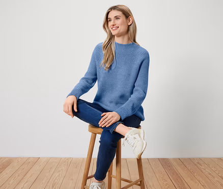 Une femme est assise sur un tabouret en bois, portant un pull bleu, un jean bleu foncé et des baskets blanches.