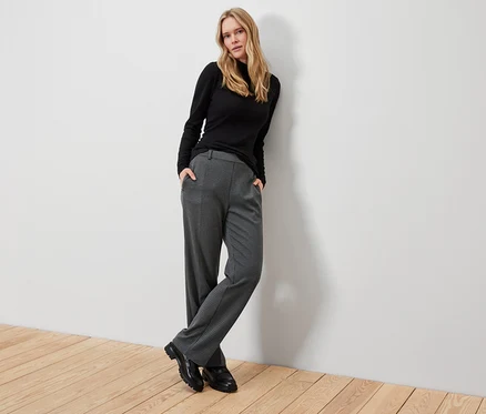 Une femme blonde pose en col roulé noir, pantalon à carreaux gris et chaussures noires.