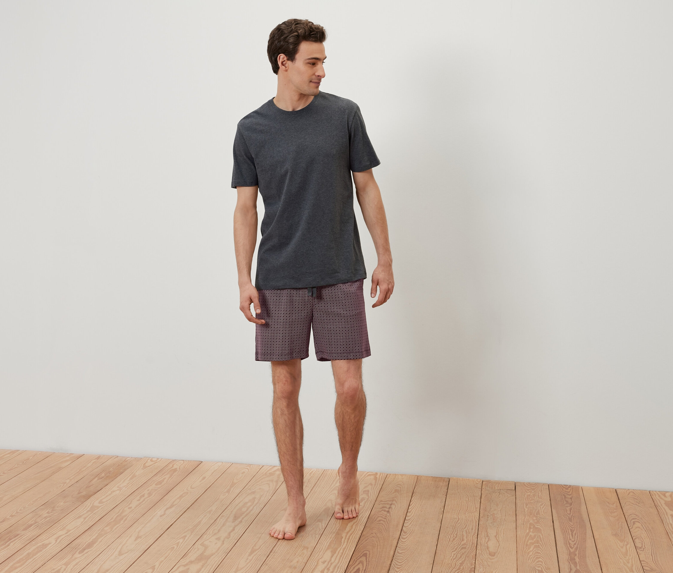 Un homme en t-shirt gris et short à motifs se tient pieds nus sur un plancher en bois devant un mur blanc.
