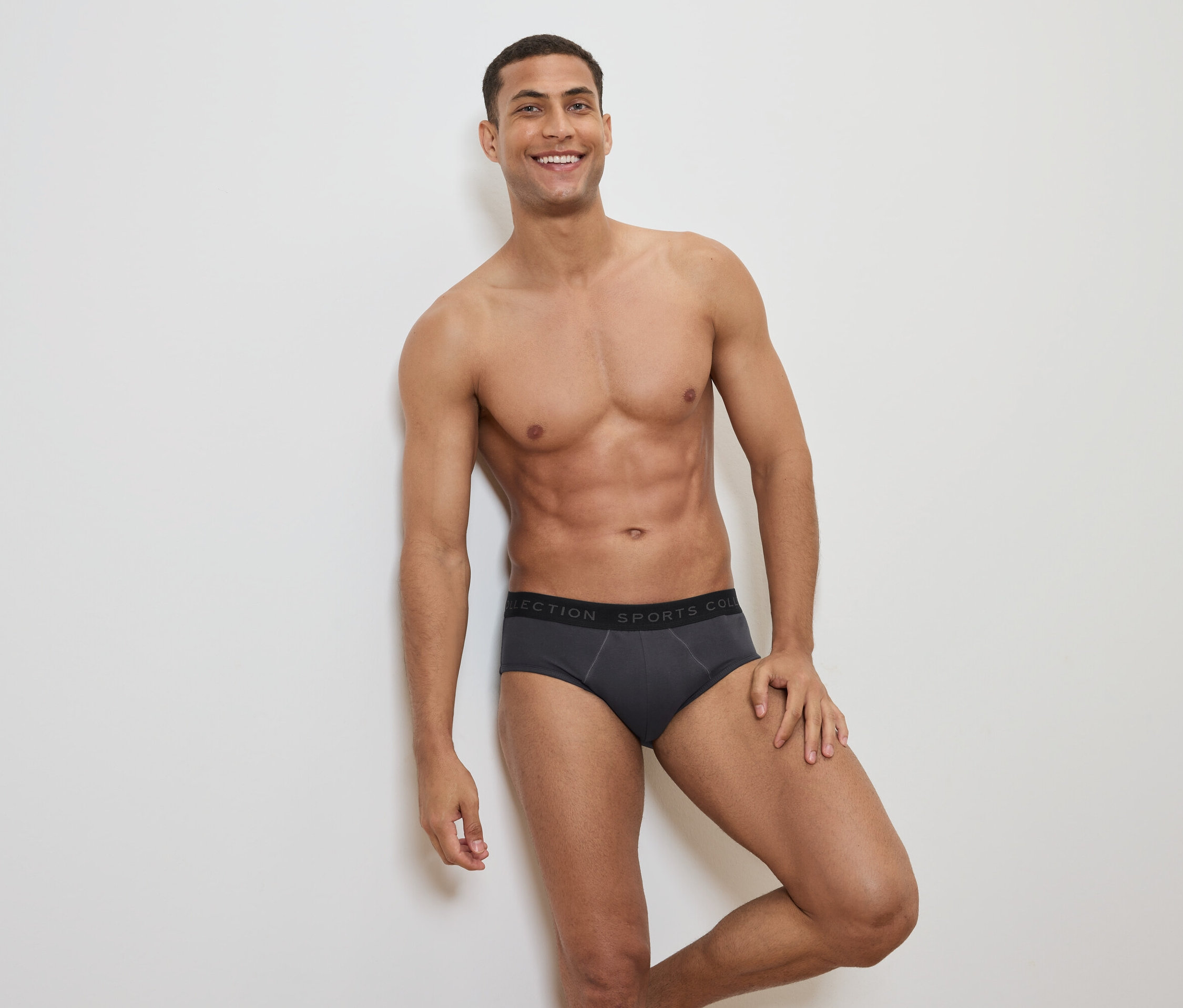 Homme souriant, torse nu, posant en slip gris avec une ceinture noire.