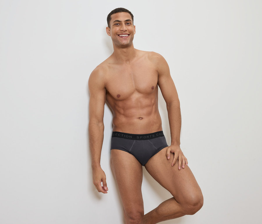 Homme souriant, torse nu, posant en slip gris avec une ceinture noire.