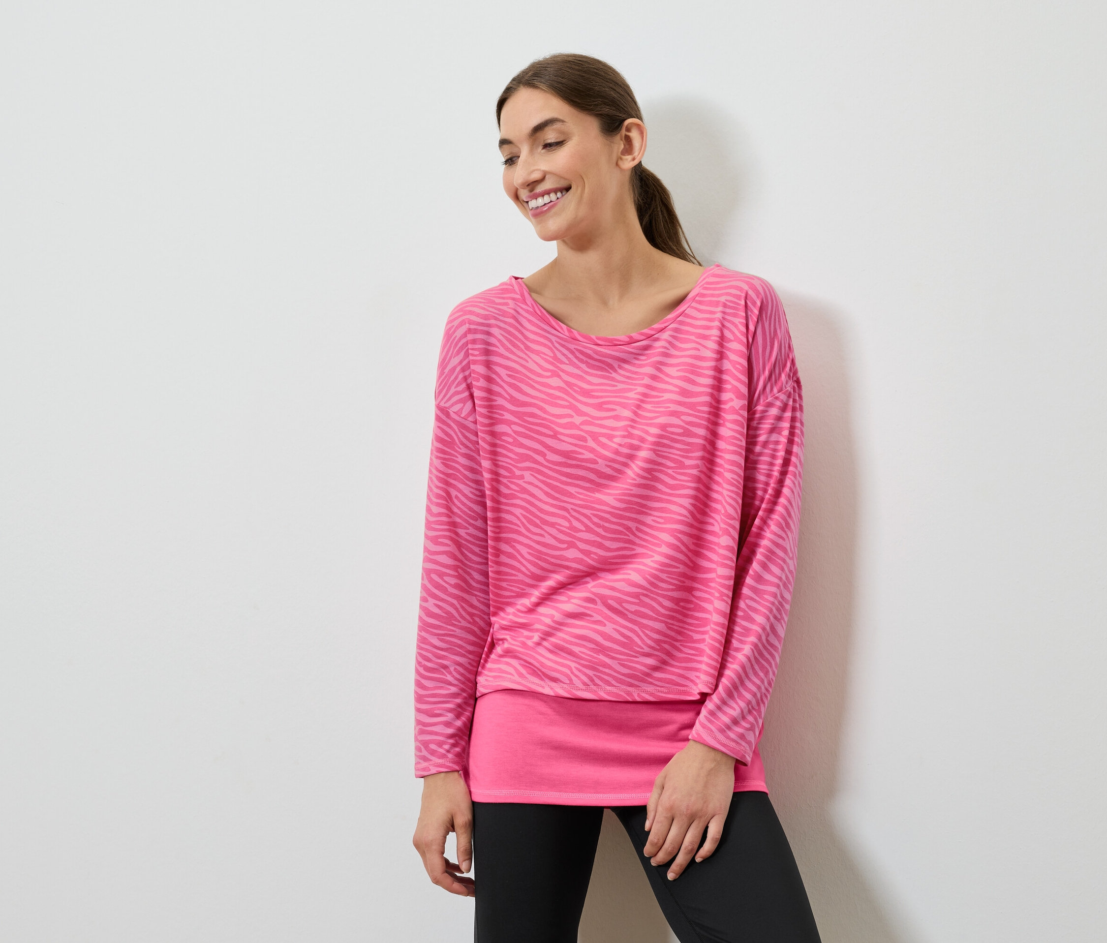 Une femme souriante pose dans un haut rose à manches longues et un legging noir.