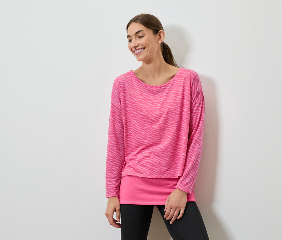 Une femme souriante pose dans un haut rose à manches longues et un legging noir.