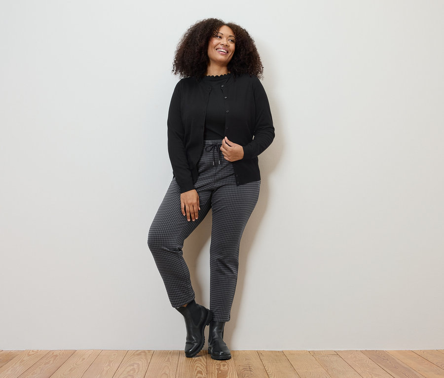 Femme posant en cardigan en maille fine, pantalon de jogging avec jacquard pied-de-poule et t-shirt noir à bordure ondulée.