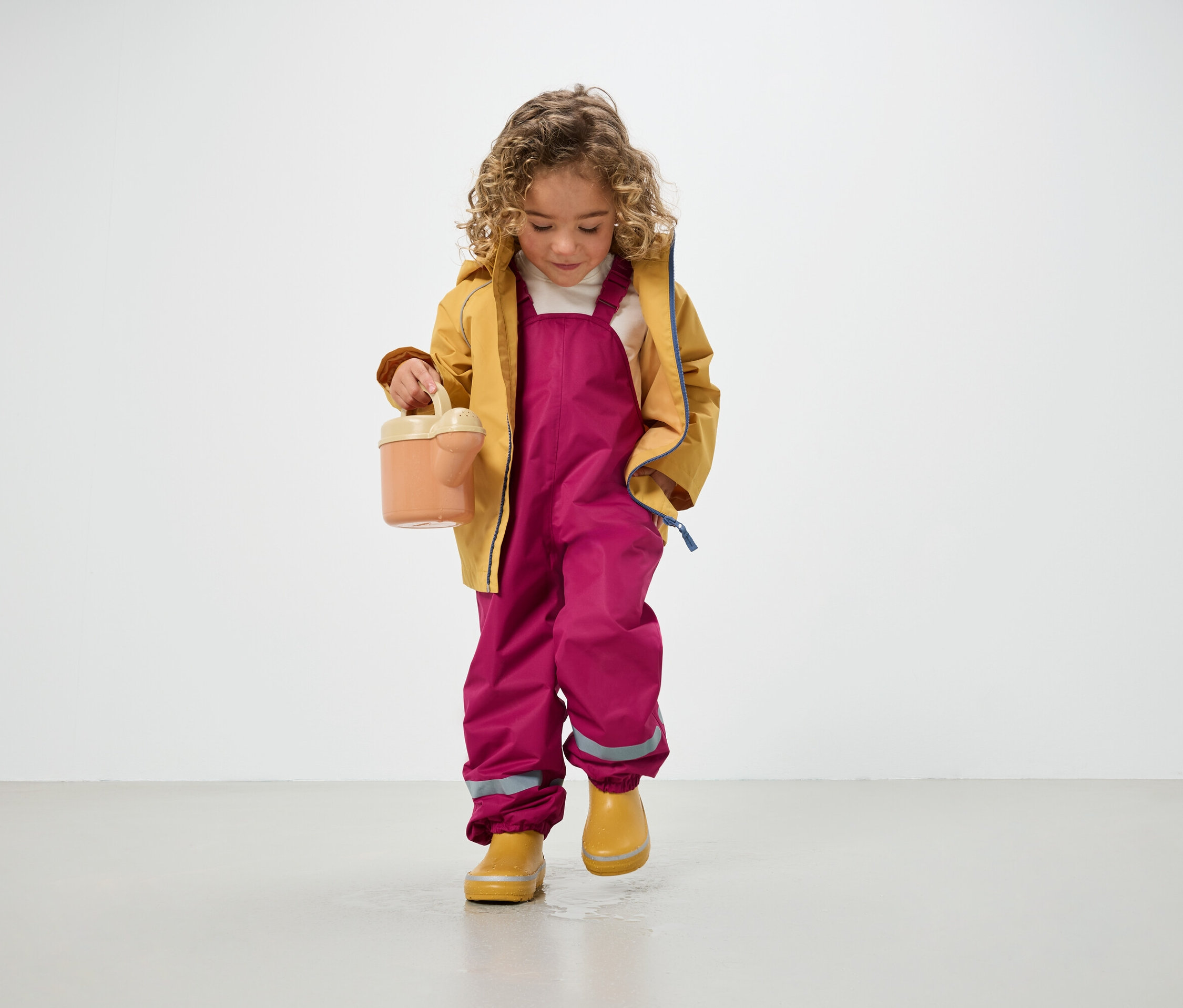 Un enfant bouclé vêtu d'une veste jaune, d'un pantalon rose et de bottes jaunes tient un arrosoir et se tient debout sur le sol.