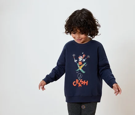 Un enfant aux cheveux bouclés foncés porte un sweat-shirt bleu marine avec une image de Mickey Mouse faisant du ski.