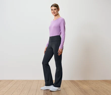 Une femme pose de profil dans un haut lilas à manches longues et un pantalon noir à taille haute.