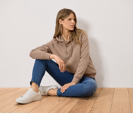 Une femme est assise sur un plancher en bois, portant un sweat à capuche marron, un jean bleu et des baskets blanches.