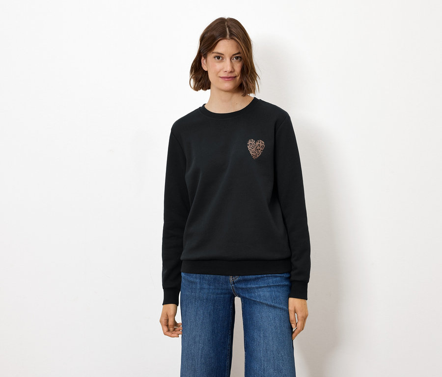 Une femme debout portant un pull noir avec un motif de cœur et un jean.