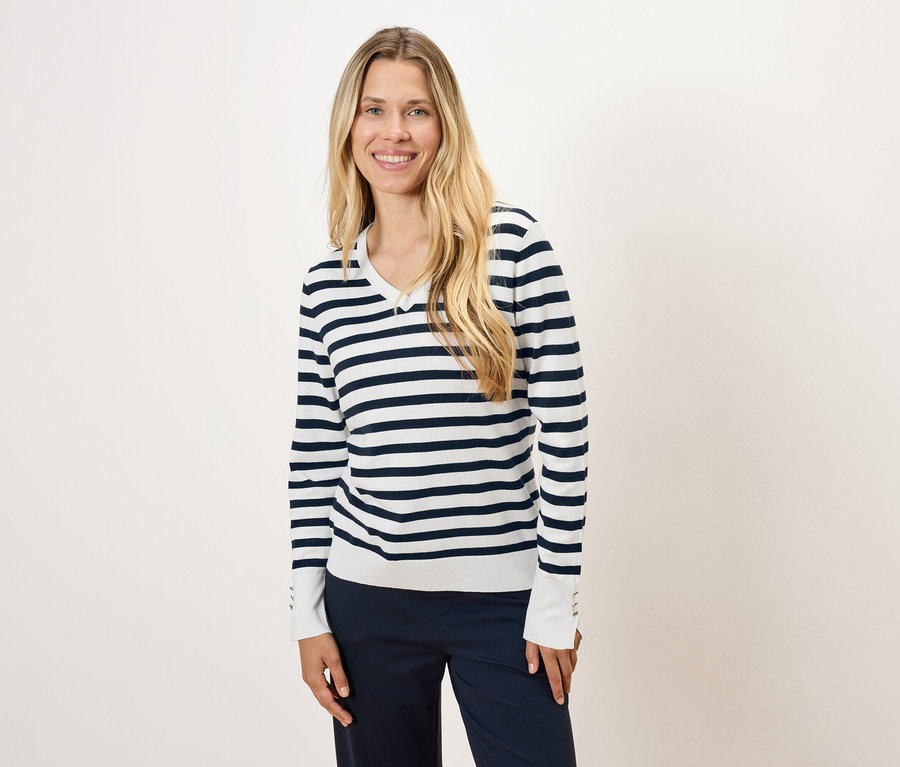 Une femme aux longs cheveux blonds porte un pull rayé et un pantalon bleu marine.