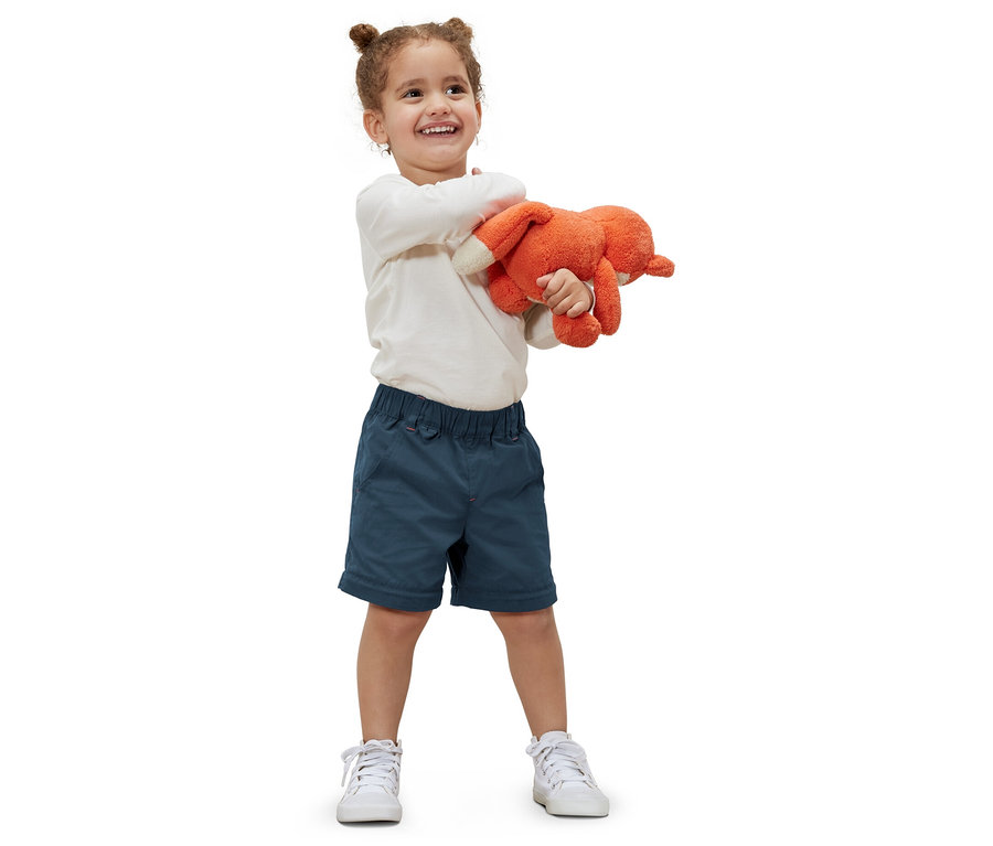 Une petite fille souriante tient une peluche orange et porte un pantalon fonctionnel pour tout-petit et des baskets blanches.