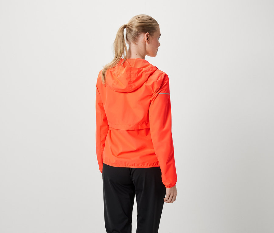 Une femme blonde portant une veste orange et un pantalon noir, les cheveux attachés en queue de cheval.