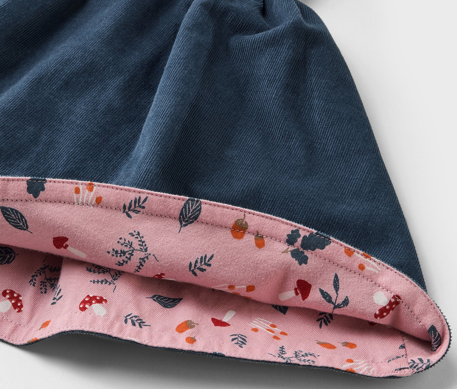Détail d'une jupe réversible en velours pour enfant, avec un côté extérieur bleu et un côté intérieur rose imprimé de feuilles et de champignons.