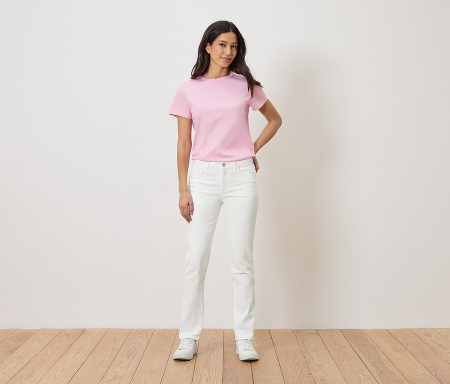 Une femme se tient devant un mur blanc, portant un t-shirt rose, un jean blanc et des baskets blanches.