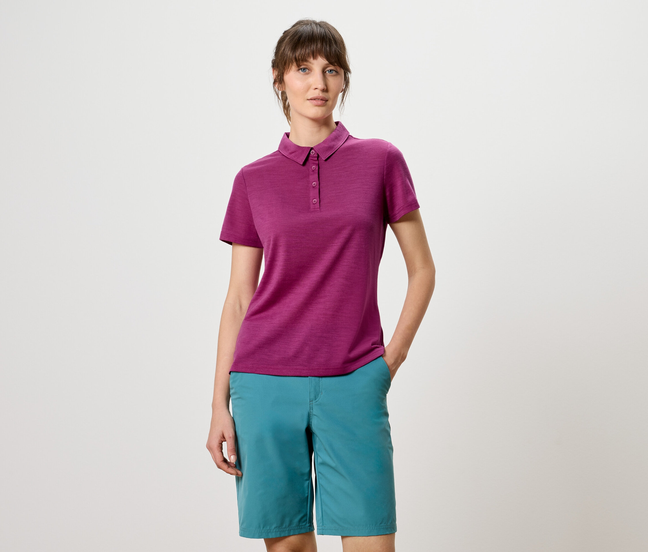 Une femme portant un polo bordeaux et un short turquoise pose devant un fond blanc.