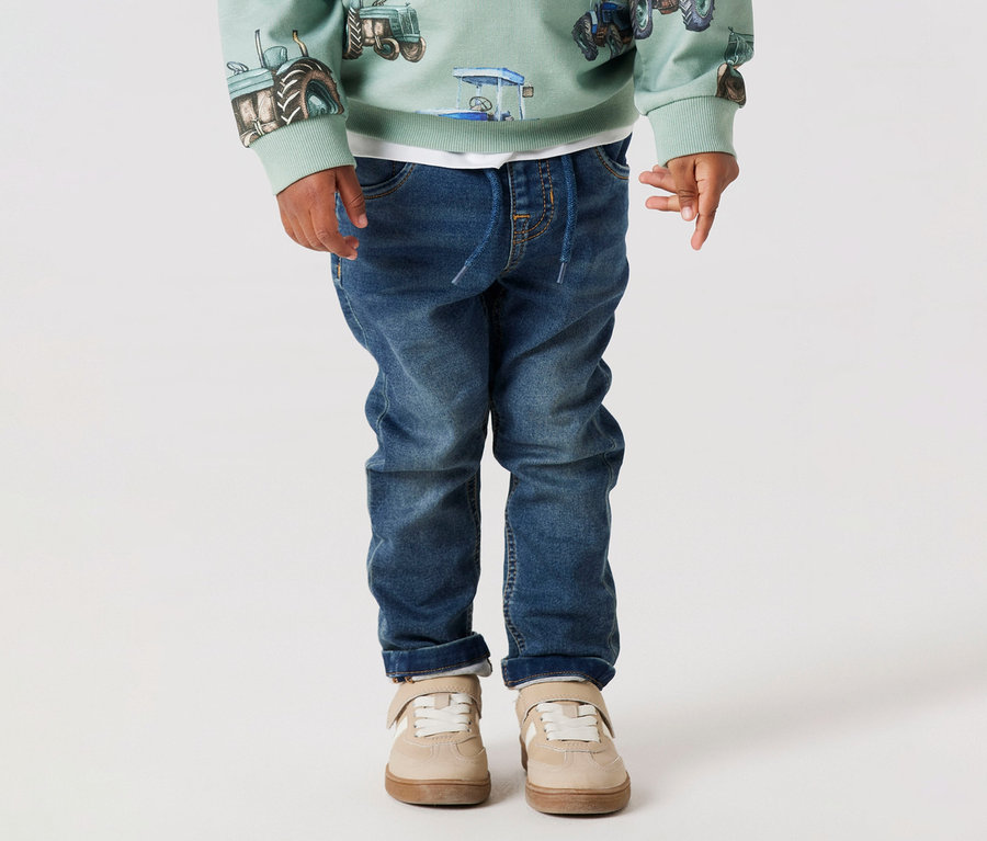 Un enfant porte un jean en sweat NAME IT, un sweat-shirt vert clair avec un motif de tracteur et des chaussures beiges sur fond blanc.