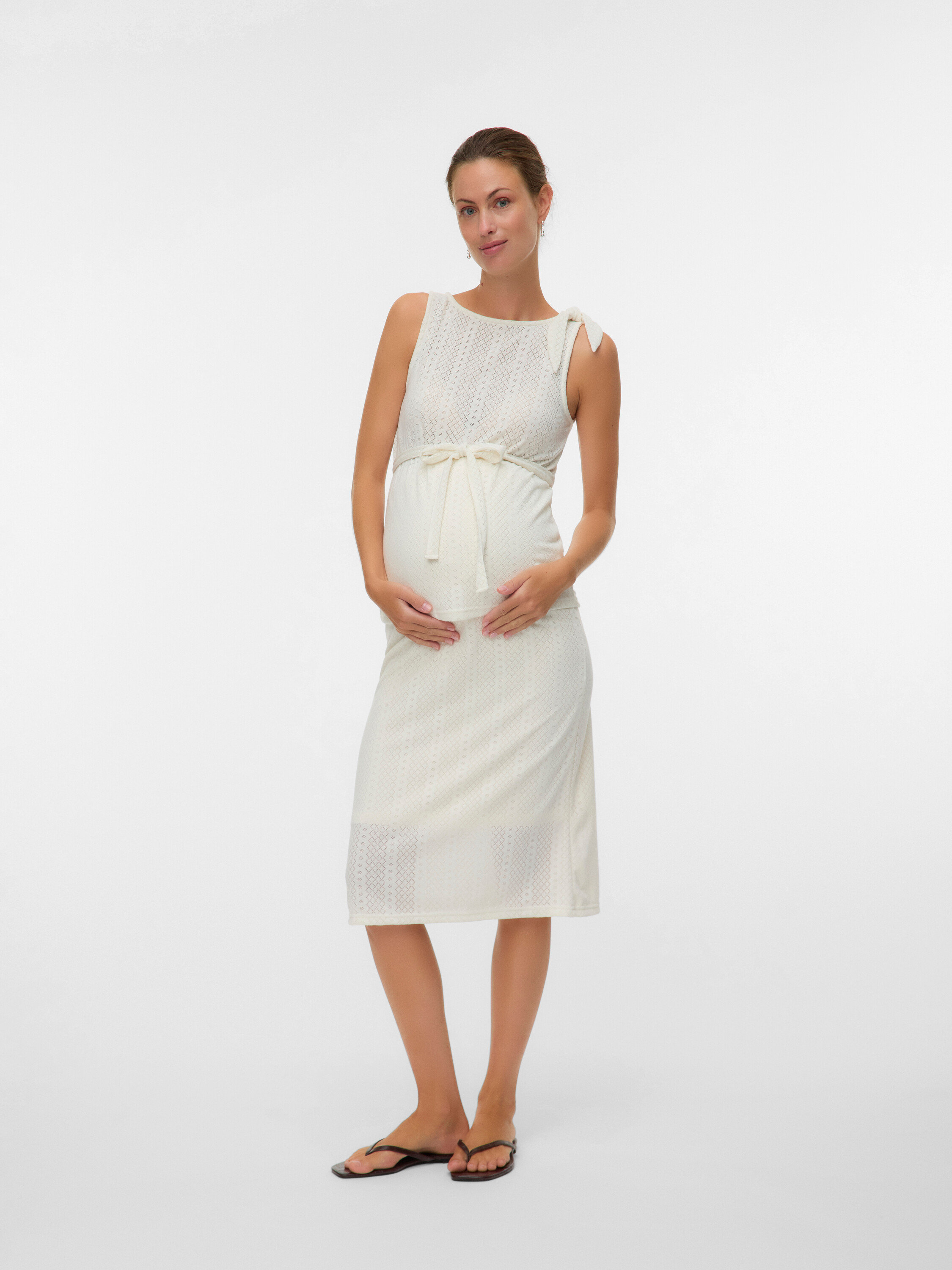 Une femme enceinte porte une robe blanche sans manches avec une ceinture à la taille et des tongs.