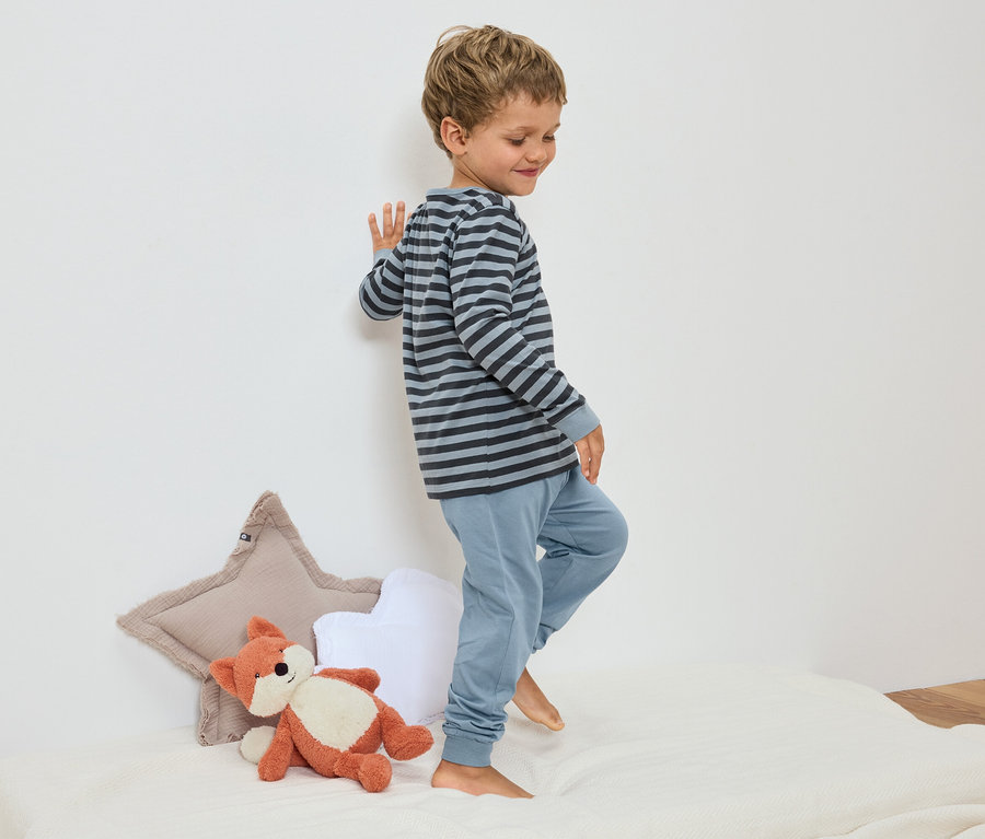 Un petit garçon en pyjama rayé se tient sur un lit à côté d'une peluche de renard et d'oreillers en forme d'étoile.