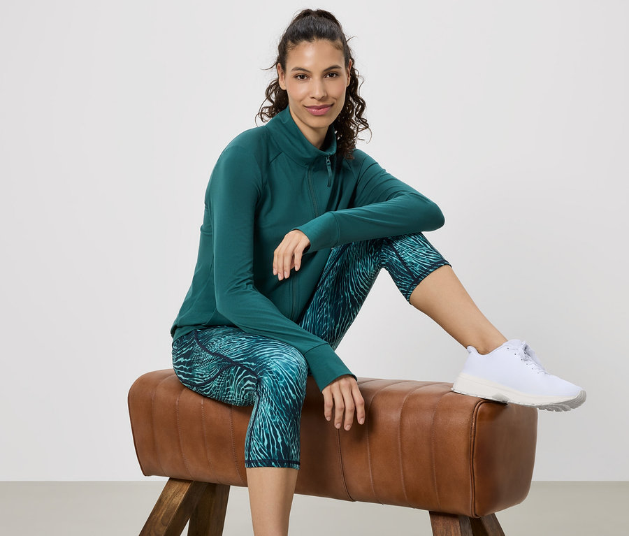 Femme assise sur un cheval d'arçons, portant une veste de sport verte, un legging à motifs et des baskets blanches.