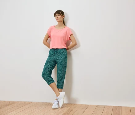 Femme adossée à un mur, portant un t-shirt corail, un pantalon capri vert à motifs de feuilles et des baskets blanches.