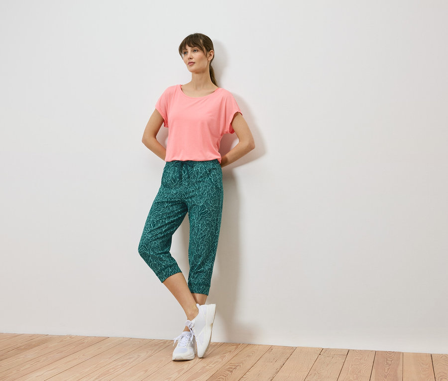 Femme adossée à un mur, portant un t-shirt corail, un pantalon capri vert à motifs de feuilles et des baskets blanches.